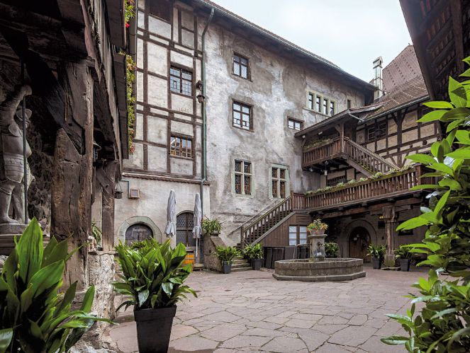 Ein historischer Steinhof mit Topfpflanzen, einem zentralen Brunnen, Holzbalkonen und Treppen, umgeben von Fachwerkwänden und Fenstern.