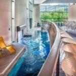 Innenpoolbereich mit einer Reihe gelber Liegestühle auf der linken Seite, einem geschwungenen Steg über dem Pool und großen Fenstern mit Blick auf das Grün draußen.