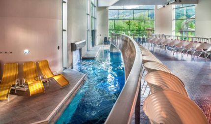 Innenpoolbereich mit einer Reihe gelber Liegestühle auf der linken Seite, einem geschwungenen Steg über dem Pool und großen Fenstern mit Blick auf das Grün draußen.