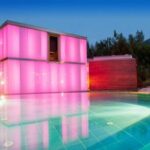 Ein modernes Gebäude mit leuchtend rosa beleuchteten Wänden steht neben einem runden, türkisfarbenen Swimmingpool, umgeben von Grün und Bäumen in der Abenddämmerung.