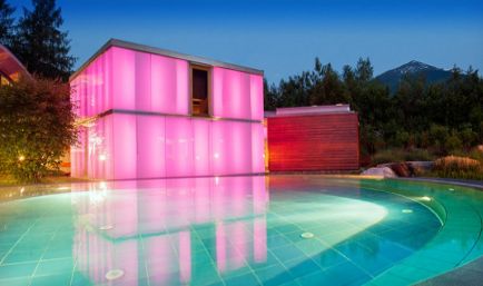 Ein modernes Gebäude mit leuchtend rosa beleuchteten Wänden steht neben einem runden, türkisfarbenen Swimmingpool, umgeben von Grün und Bäumen in der Abenddämmerung.