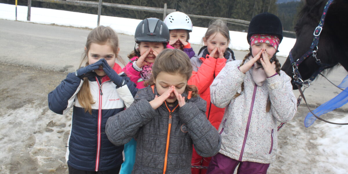 Sechs Kinder in Winterjacken machen lustige Grimassen im Freien in der Nähe einer verschneiten Straße, wobei ein Pferd auf der rechten Seite des Bildes teilweise sichtbar ist.
