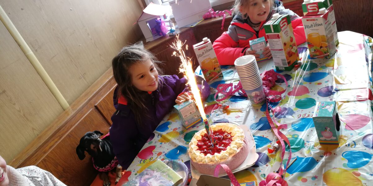 Zwei Kinder sitzen an einem Geburtstagstisch mit einer Torte mit Wunderkerzen, bunten Dekorationen, Saftkartons und einem kleinen Hund in der Nähe.