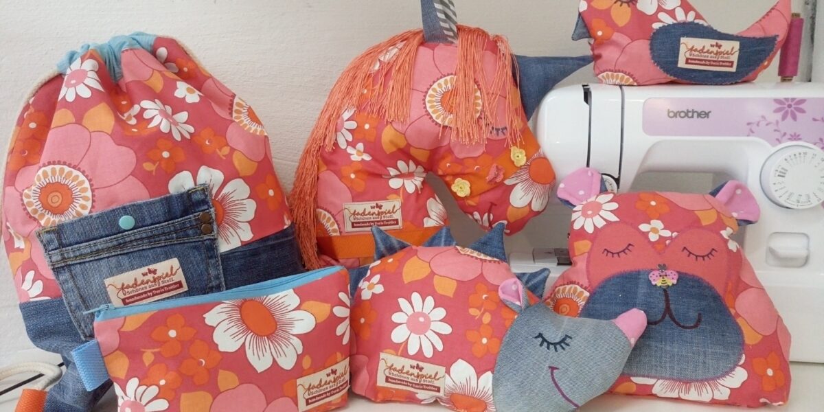 Eine Sammlung von handgefertigten Stoffartikeln mit rosa und orangefarbenen Blumenmustern, darunter Kissen in Tierform, Beutel, eine Tasche mit Kordelzug und eine Nähmaschine im Hintergrund.