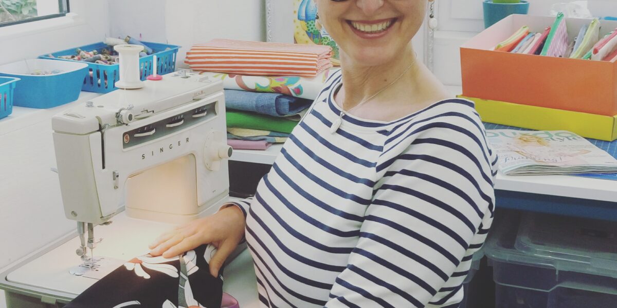 Frau mit kurzen braunen Haaren und Brille lächelt, während sie eine Nähmaschine auf einem geblümten Stoff in einem hellen, organisierten Arbeitsbereich bedient.