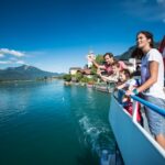 Eine Familie steht auf einem Bootsdeck und blickt auf einen See mit Bergen und Dorfgebäuden im Hintergrund unter einem klaren blauen Himmel.