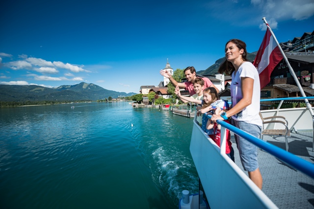 Eine Familie steht auf einem Bootsdeck und blickt auf einen See mit Bergen und Dorfgebäuden im Hintergrund unter einem klaren blauen Himmel.