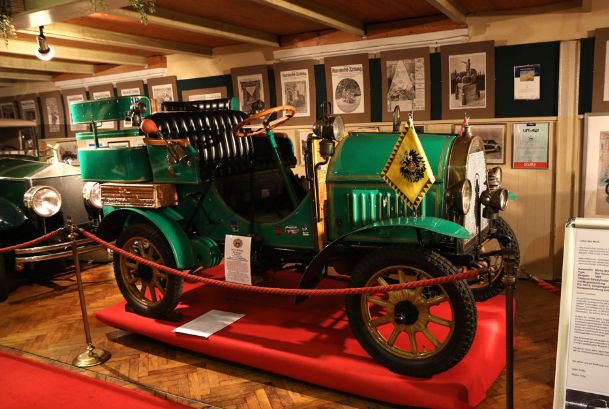 Ein grüner Oldtimer steht auf einer roten Plattform in einem Museum, umgeben von Informationsschildern, gerahmten Dokumenten und einem roten Absperrseil.