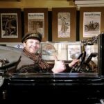 Ein Mann mit Mütze und Schal sitzt auf dem Fahrersitz eines Oldtimers, lächelt und zeigt den Daumen nach oben; an der Wand hinter ihm hängen alte Autoposter.