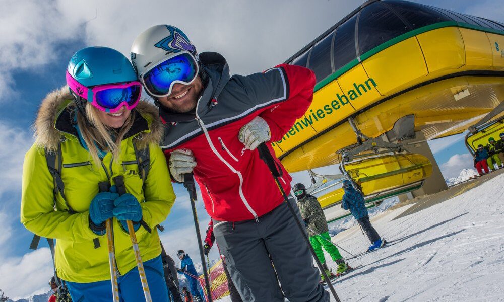 Zwei Skifahrer in hellen Jacken posieren mit ihrer Skiausrüstung vor einem Skilift auf einem verschneiten Berg mit anderen Skifahrern in der Nähe.