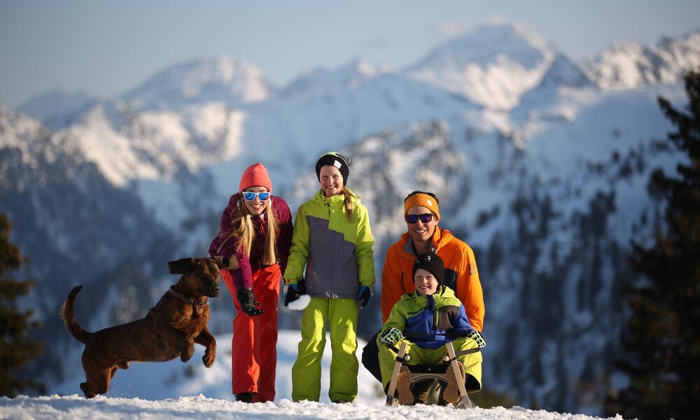 Vier Personen und ein Hund posieren auf einem verschneiten Berg mit schneebedeckten Gipfeln im Hintergrund; eine Person sitzt auf einem Schlitten, während die anderen in der Nähe stehen oder hocken.