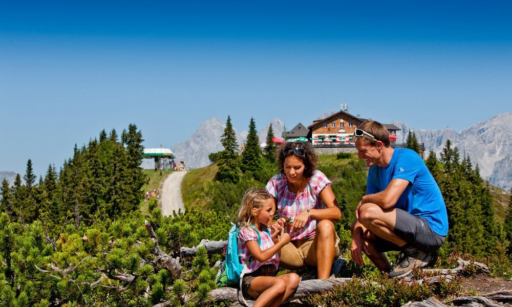 Eine Familie sitzt auf einem Bergpfad, umgeben von Grün, mit einer Hütte und Bergen im Hintergrund unter einem klaren blauen Himmel.