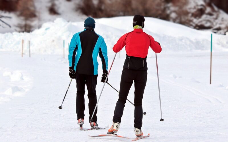 Zwei Skilangläufer auf einer verschneiten Loipe, ein weiterer Skifahrer ist im Hintergrund in der Nähe einer Schneewehe zu sehen.
