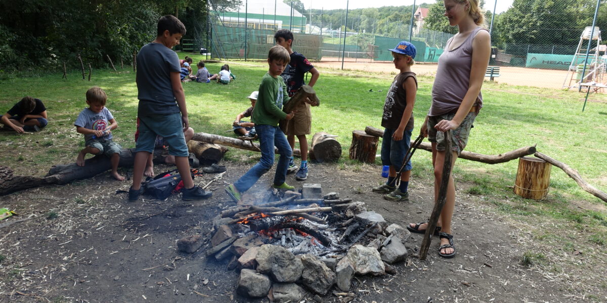 Eine Gruppe von Kindern und ein Erwachsener stehen um ein Lagerfeuer in einem grasbewachsenen Außenbereich mit Spielplatz und Sportplätzen im Hintergrund.