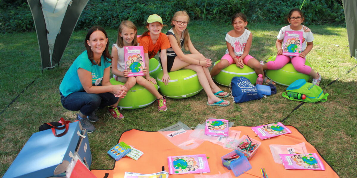 Eine Gruppe von Kindern und ein Erwachsener sitzen im Freien auf grünen Kissen unter einem Vordach, halten Activity-Bücher in der Hand und haben ihr Malmaterial auf Matten vor sich ausgebreitet.