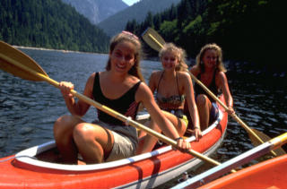 Drei Personen paddeln an einem sonnigen Tag in einem roten Kanu auf einem ruhigen See, der von bewaldeten Bergen umgeben ist.