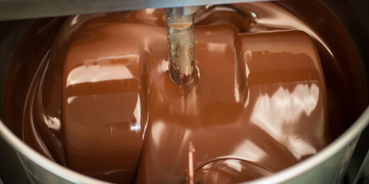 Industrielle Maschine, die geschmolzene Schokolade in einer großen Schüssel aus rostfreiem Stahl mischt, wobei die Schokolade um rotierende Schaufeln wirbelt.