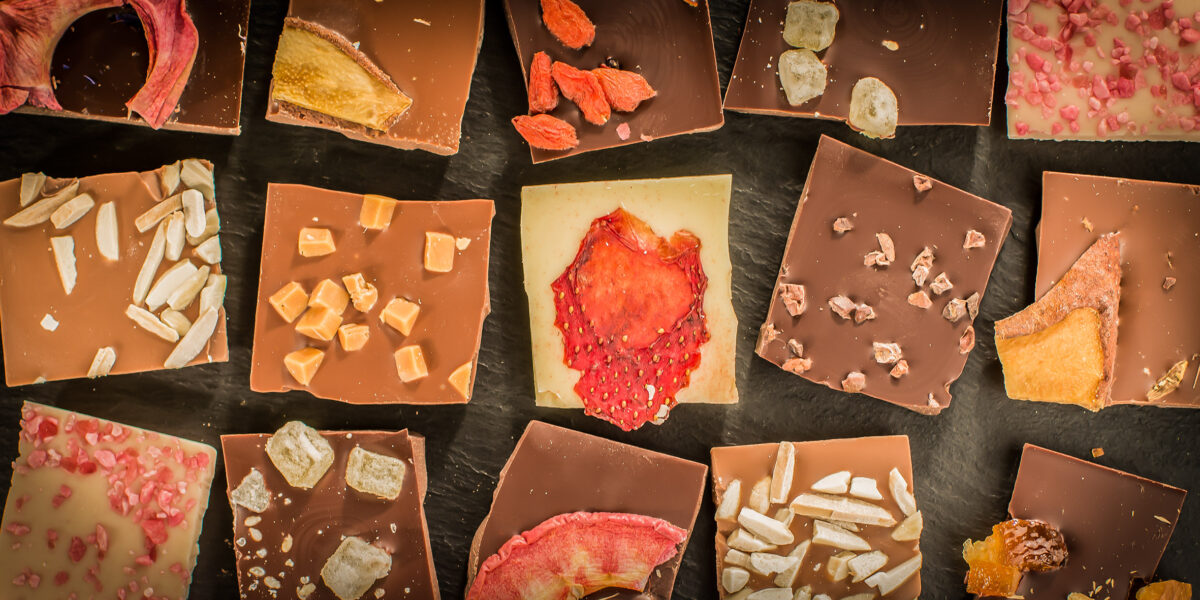 Gemischte Schokoladenquadrate mit verschiedenen Belägen wie Trockenfrüchten, Nüssen, Samen und Bonbons auf einer dunklen Oberfläche.