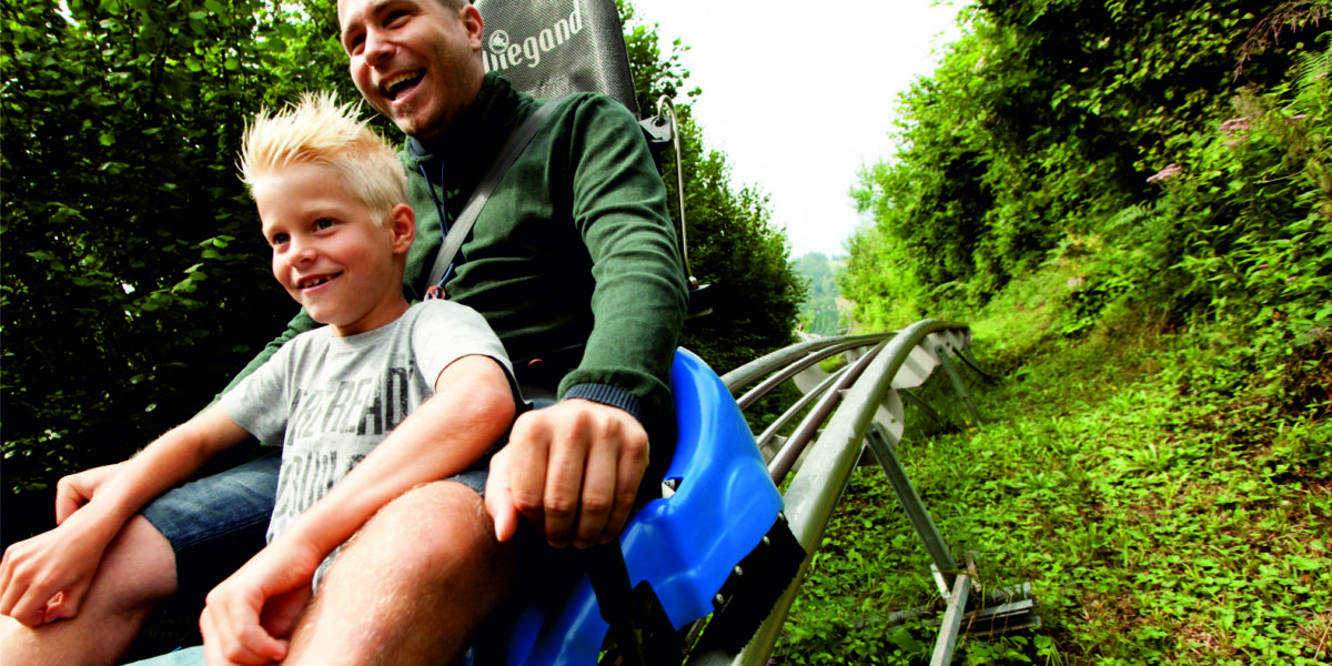 Ein Erwachsener und ein Kind fahren gemeinsam auf einer blauen Alpenachterbahn, die von grünen Bäumen und Gras umgeben ist, auf einer Abfahrtsstrecke.