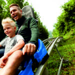 Ein Erwachsener und ein Kind fahren gemeinsam auf einer blauen Alpenachterbahn, die von grünen Bäumen und Gras umgeben ist, auf einer Abfahrtsstrecke.