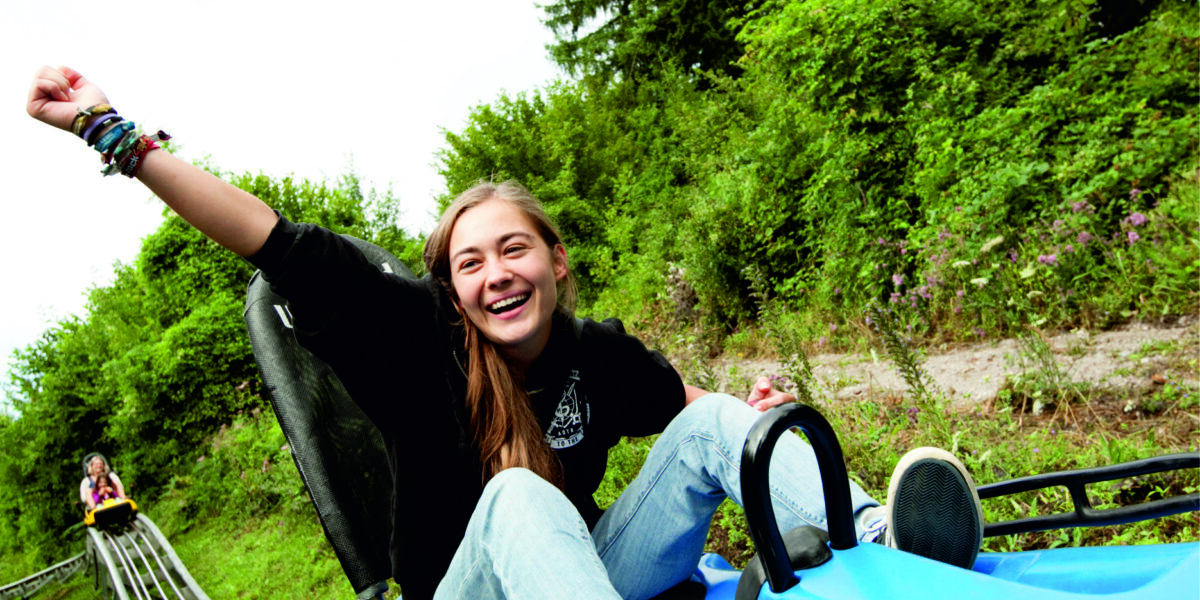 Eine junge Person fährt im Freien auf einem blauen Alpine Coaster, lächelt und hebt einen Arm, während im Hintergrund eine Grünfläche und ein anderer Fahrer zu sehen sind.