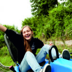 Eine junge Person fährt im Freien auf einem blauen Alpine Coaster, lächelt und hebt einen Arm, während im Hintergrund eine Grünfläche und ein anderer Fahrer zu sehen sind.