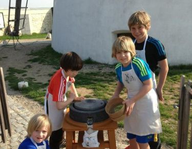 Vier Kinder in Schürzen stehen im Freien um eine Steinmühle herum. Ein Kind mahlt Getreide, andere schauen zu oder helfen mit. Im Hintergrund ist ein weißes Gebäude zu sehen.