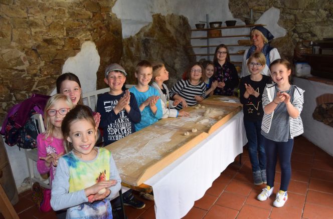 Eine Gruppe Kinder und ein Erwachsener stehen lächelnd um einen langen, mit Mehl bedeckten Holztisch herum und formen Teig in einem rustikalen Raum mit Steinwänden und Regalen.