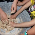 Drei Personen kneten Teig in einer großen Schüssel, wobei das Kind rechts ein gelbes Hemd und ein blaues Armband trägt.