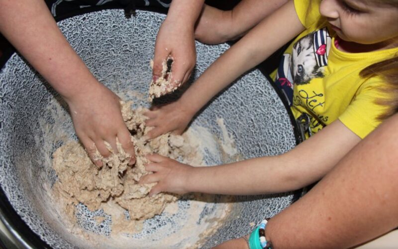 Drei Personen kneten Teig in einer großen Schüssel, wobei das Kind rechts ein gelbes Hemd und ein blaues Armband trägt.