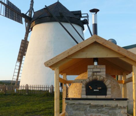 Im Hintergrund steht eine weiße Windmühle, im Vordergrund ein holzbefeuerter Außenofen unter einem Holzunterstand.