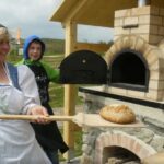 Eine Frau in traditioneller Kleidung hält einen Holzschieber mit einem Laib Brot neben einem Steinofen im Freien, neben ihr steht ein Junge.
