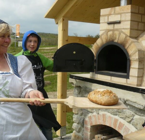 Eine Frau in traditioneller Kleidung hält einen Holzschieber mit einem Laib Brot neben einem Steinofen im Freien, neben ihr steht ein Junge.