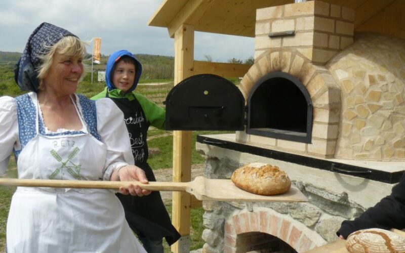 Eine Frau in traditioneller Kleidung hält einen Holzschieber mit einem Laib Brot neben einem Steinofen im Freien, neben ihr steht ein Junge.