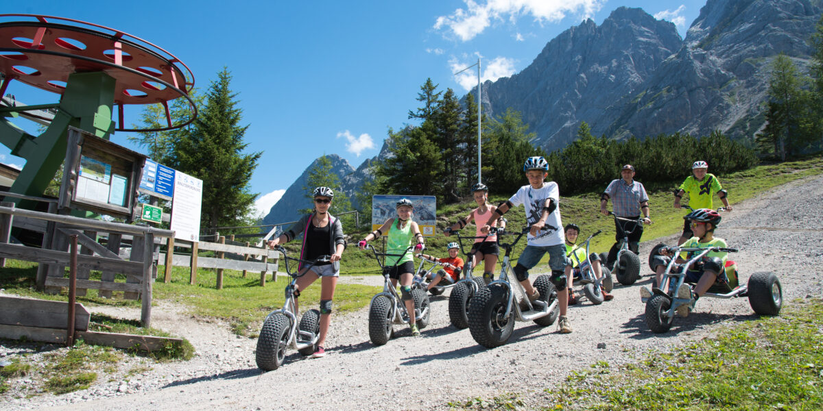 Eine Gruppe von Kindern und Erwachsenen fährt auf Mountainbikes einen Schotterweg in einer bergigen, bewaldeten Gegend unter blauem Himmel hinunter.