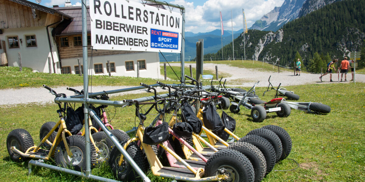 Mehrere gelbe Mountaincarts sind an einer "Rollerstation" in einer malerischen Alpenlandschaft mit Bergen und Menschen im Hintergrund abgestellt.