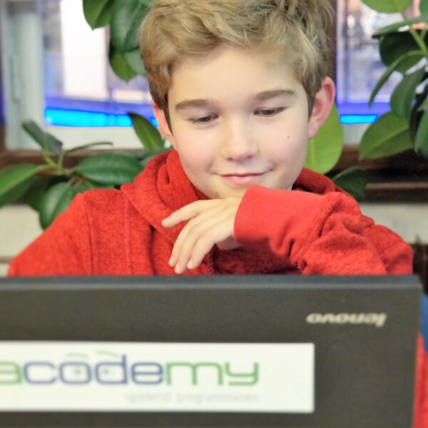 Ein junger Mensch in einem roten Kapuzenpulli sitzt vor einem Laptop mit einem „Acodemy“-Aufkleber, blickt auf den Bildschirm und stützt sein Kinn auf die Hand.