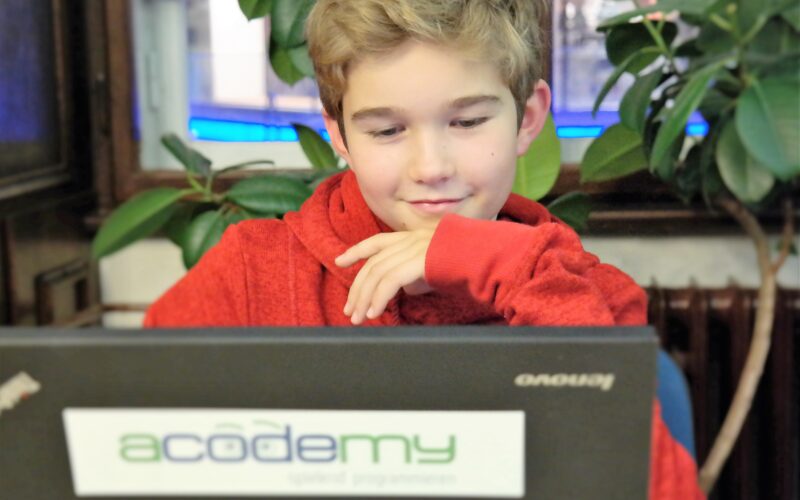 Ein junger Mensch in einem roten Kapuzenpulli sitzt vor einem Laptop mit einem „Acodemy“-Aufkleber, blickt auf den Bildschirm und stützt sein Kinn auf die Hand.