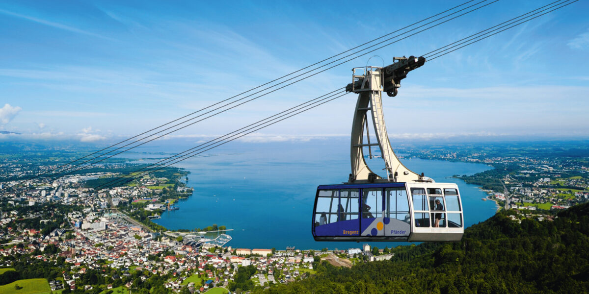 Eine Seilbahn fährt über einen grünen Hügel, von dem aus unter einem klaren blauen Himmel ein großer See, eine Stadt und eine Landschaft in der Ferne zu sehen sind.