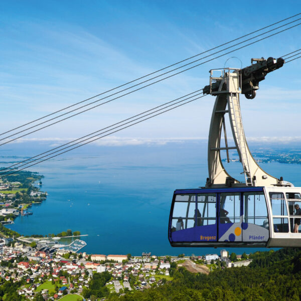 Eine Seilbahn fährt über einen grünen Hügel, von dem aus unter einem klaren blauen Himmel ein großer See, eine Stadt und eine Landschaft in der Ferne zu sehen sind.