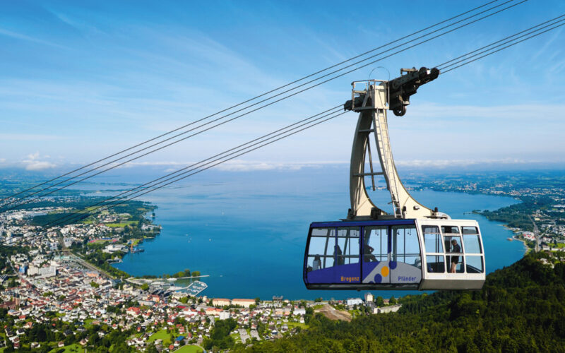 Eine Seilbahn fährt über einen grünen Hügel, von dem aus unter einem klaren blauen Himmel ein großer See, eine Stadt und eine Landschaft in der Ferne zu sehen sind.