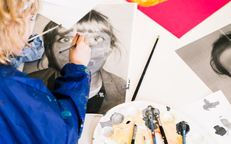 Ein Kind in einem blauen Kittel übermalt ein Schwarzweiß-Porträtfoto. Auf dem Tisch liegen Pinsel, eine Palette und Malutensilien verstreut.