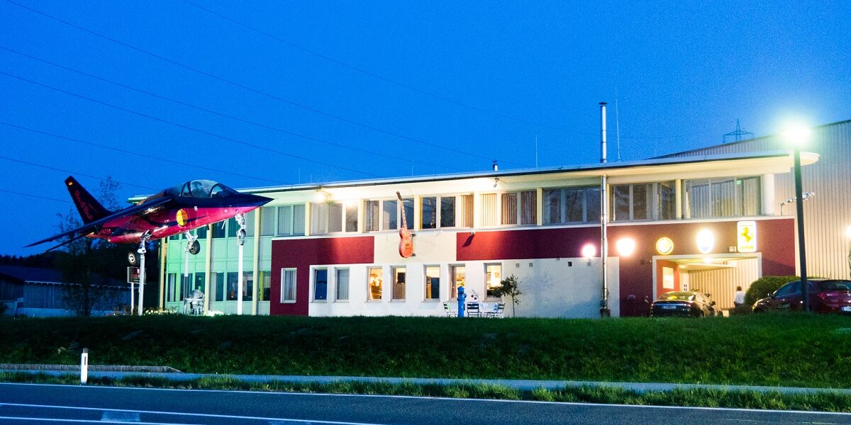 Ein kleines, modernes Gebäude ist in der Abenddämmerung beleuchtet, und ein rotes Düsenflugzeug ist an einem Mast in der Nähe einer Straße angebracht.