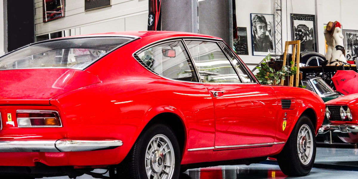 Ein klassischer roter Sportwagen mit Ferrari-Plaketten ist in einem Ausstellungsraum geparkt, umgeben von gerahmten Fotos und Postern an den Wänden.