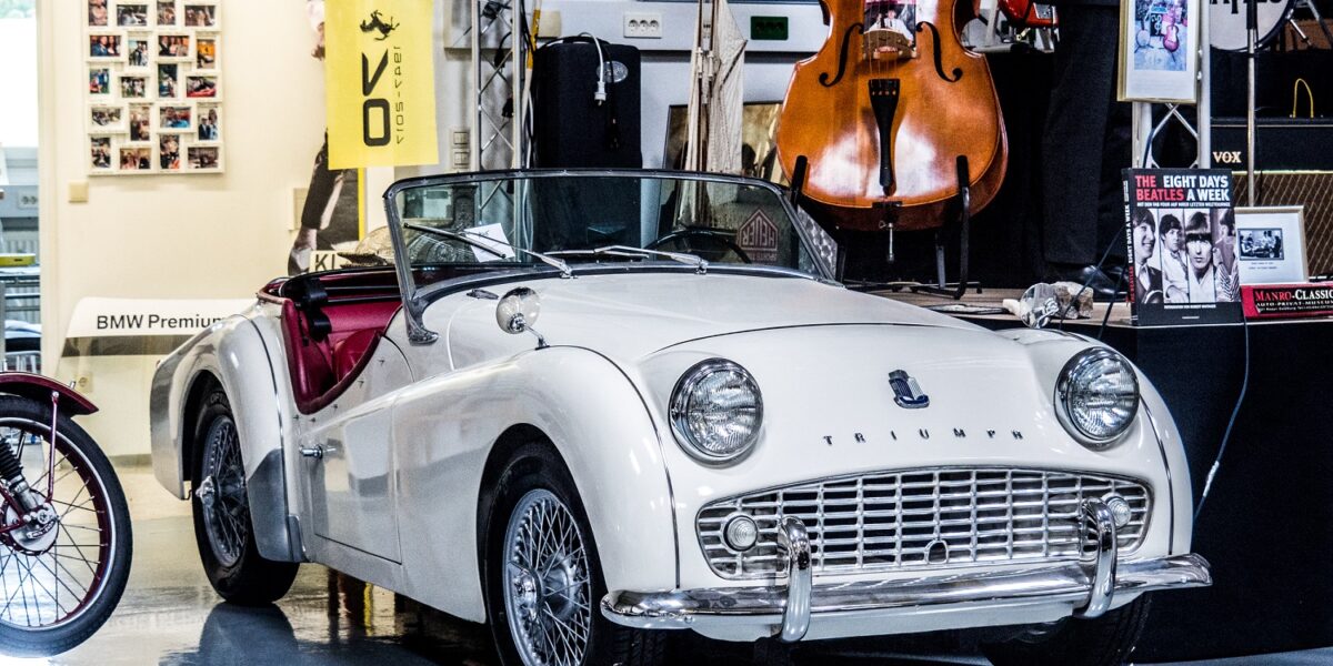 Ein weißer Triumph-Oldtimer mit Cabrio ist in einem Raum neben Musikinstrumenten, gerahmten Fotos und Beatles-Erinnerungsstücken ausgestellt.