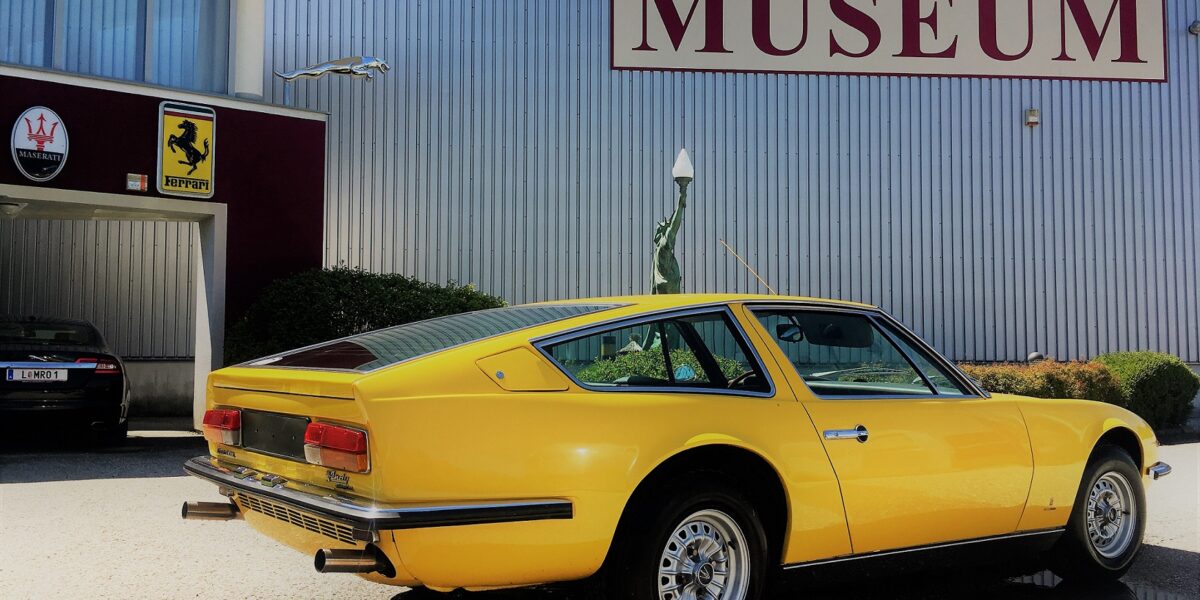 Ein gelber klassischer Sportwagen parkt vor einem Gebäude mit einem großen MUSEUM"-Schild und Automobillogos an der Wand.