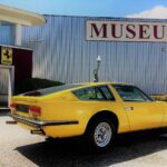 Ein gelber klassischer Sportwagen parkt vor einem Gebäude mit einem großen MUSEUM"-Schild und Automobillogos an der Wand.
