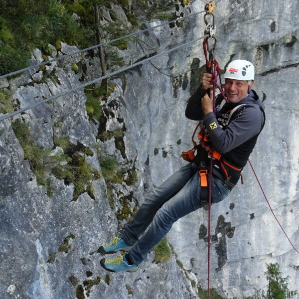 Person mit Sicherheitsausrüstung und Helm beim Ziplining über eine Felswand, im Hintergrund ist die Vegetation zu sehen.