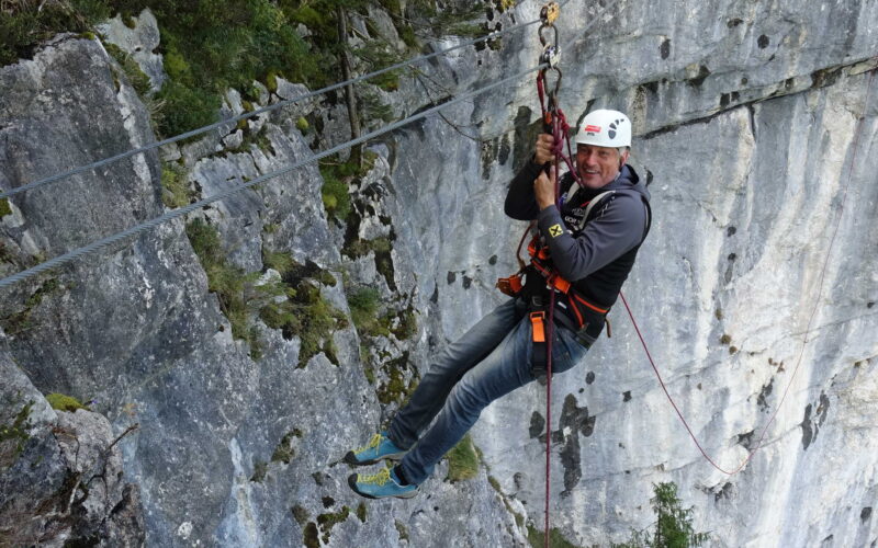 Person mit Sicherheitsausrüstung und Helm beim Ziplining über eine Felswand, im Hintergrund ist die Vegetation zu sehen.