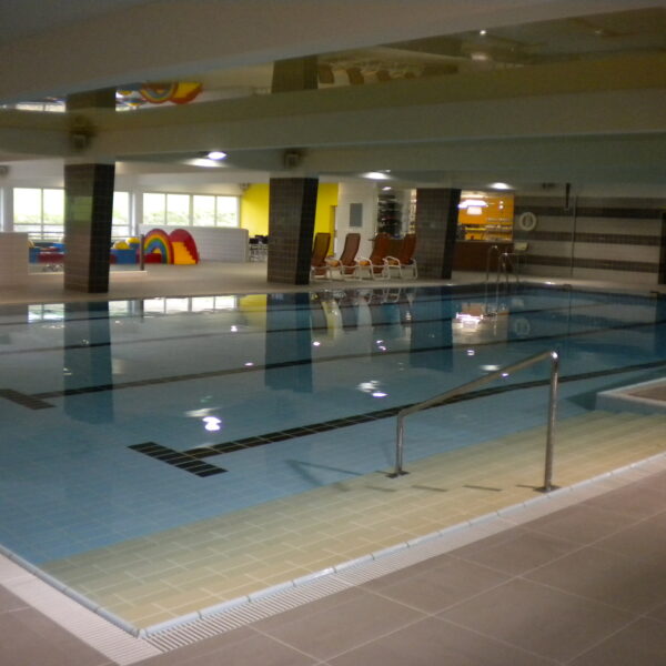 Innenpool mit klarem Wasser, Poolleiter, gefliestem Boden und Liegestühlen im Hintergrund unter fluoreszierender Beleuchtung.